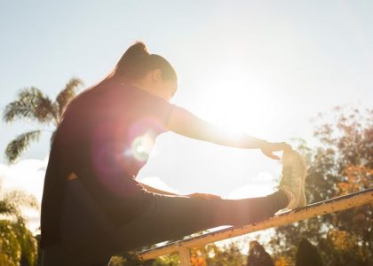 A woman stretches on a bar in a sunlit park, surrounded by trees and a clear sky.