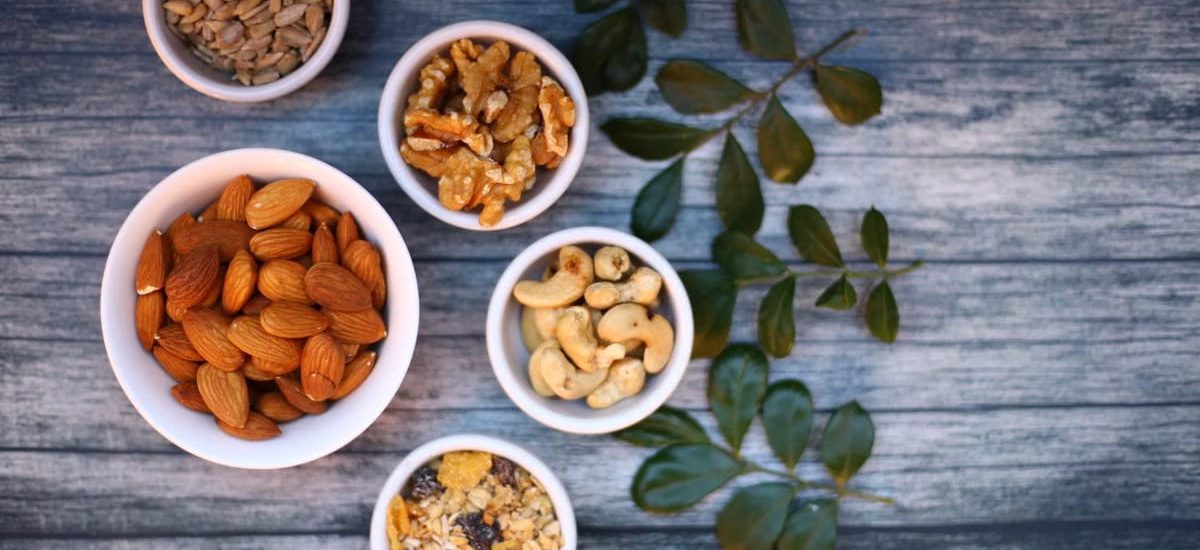 Aerial view of assorted nuts and seeds in white bowls with green leaves on a rustic wooden table.