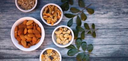 Aerial view of assorted nuts and seeds in white bowls with green leaves on a rustic wooden table.