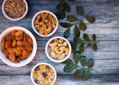Aerial view of assorted nuts and seeds in white bowls with green leaves on a rustic wooden table.